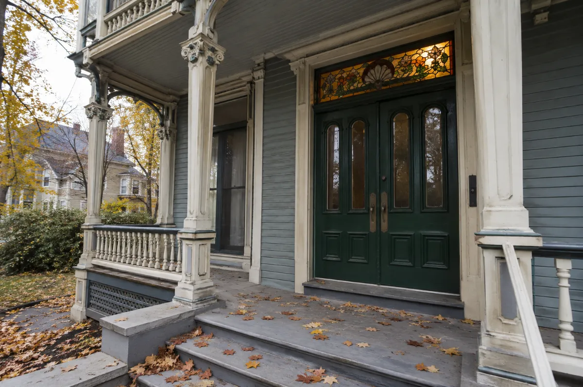 Grand Victorian double-door entry in deep green with a stained-glass transom on a Bangor mansion porch, autumn leaves on the steps