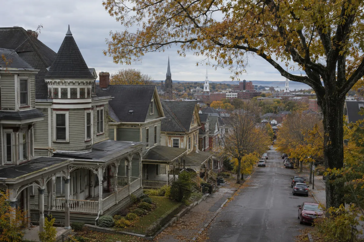 Elevated autumn view of a grand Bangor Maine residential street — lumber-era Victorian mansions with turrets and wraparound porches under towering maples, church steeples and city rooftops beyond