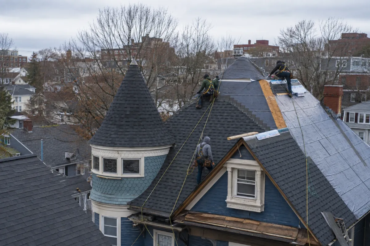 Roofing crew installing charcoal architectural shingles on a steep Bangor Victorian with a turret, city neighborhood rooftops and bare maples behind