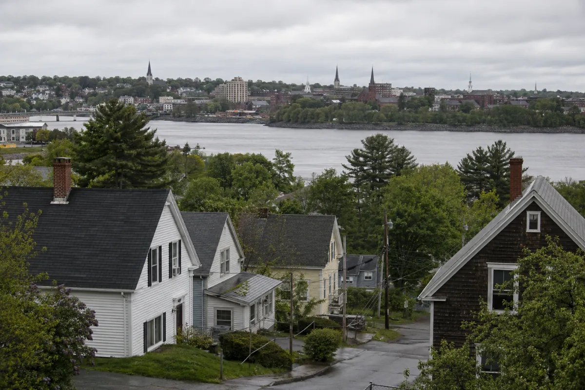 Brewer Maine hillside neighborhood — capes and New Englanders stepping down toward the Penobscot River, Bangor's steeples and rooftops across the water