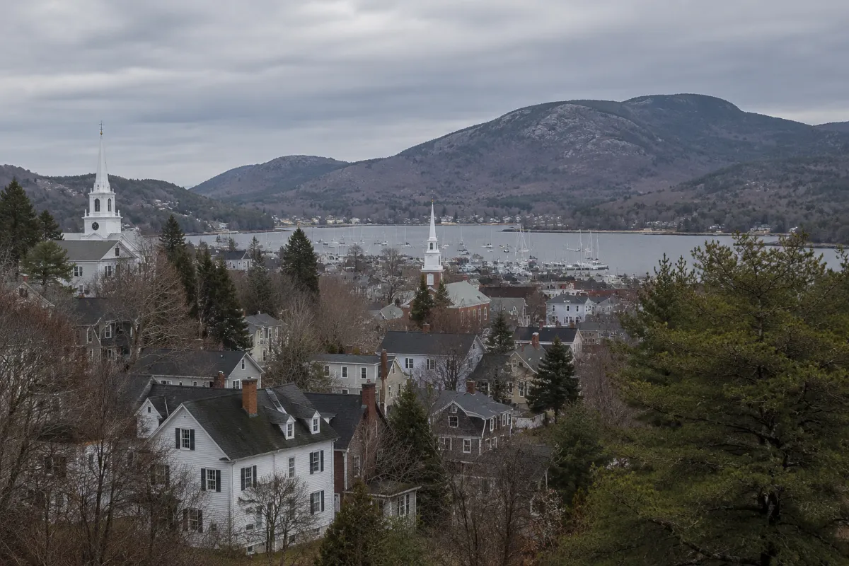 Elevated view of Camden Maine village — white sea captains' homes and church steeples stepping downhill to a harbor full of schooner masts, Mount Battie and the Camden Hills rising behind