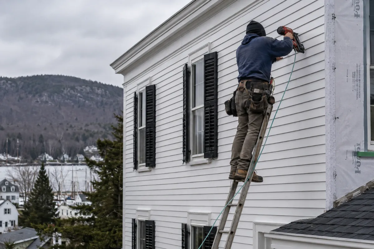 Installer nailing crisp white clapboard siding on a historic Camden colonial with black shutters, wooded Camden Hills behind