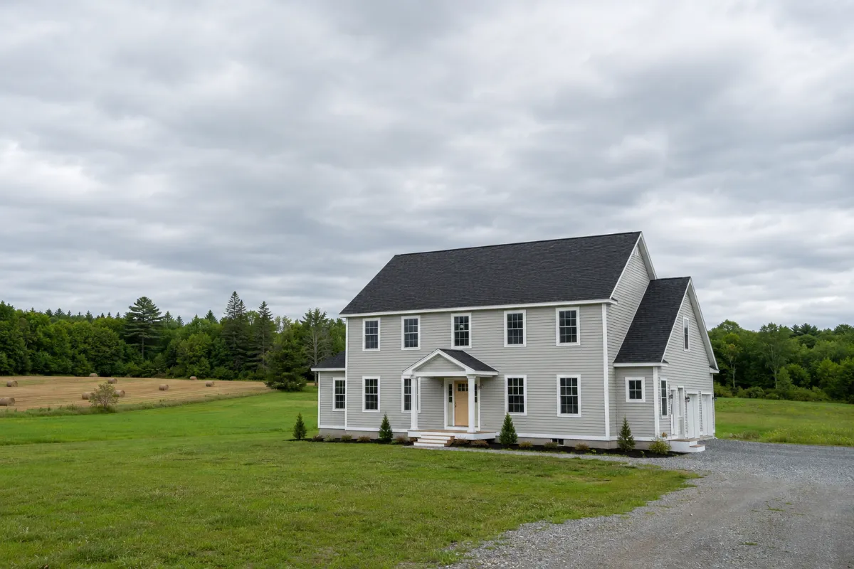 Newer Hermon Maine colonial on a big open lot — fresh architectural-shingle roof and crisp siding under a huge sky, hayfields and treeline beyond
