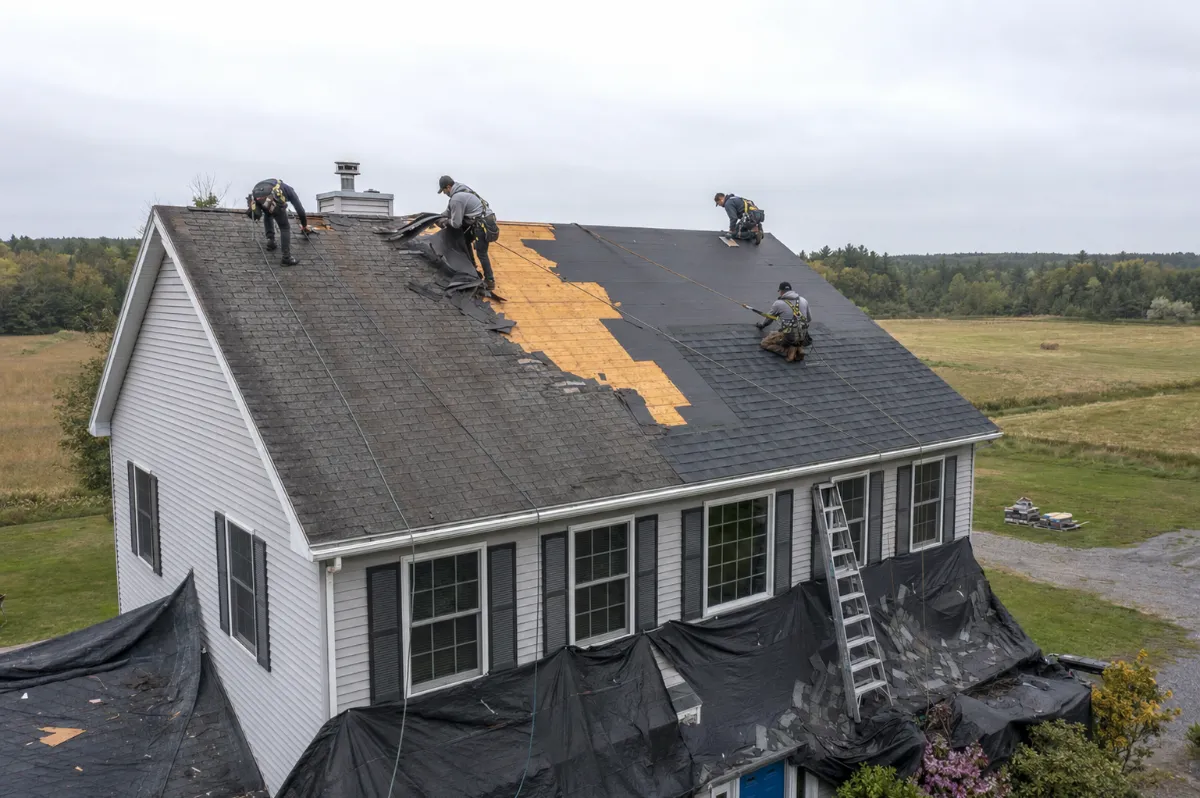Crew tearing off worn builder-grade shingles and installing architectural shingles on a 2000s Hermon colonial, open fields behind