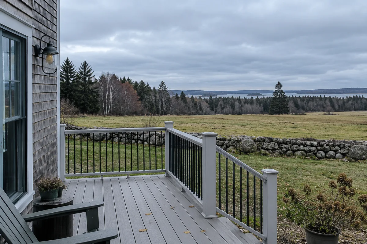 New composite deck on a rural Winterport Maine farmhouse looking across a hayfield toward the treeline, old stone wall at the field edge