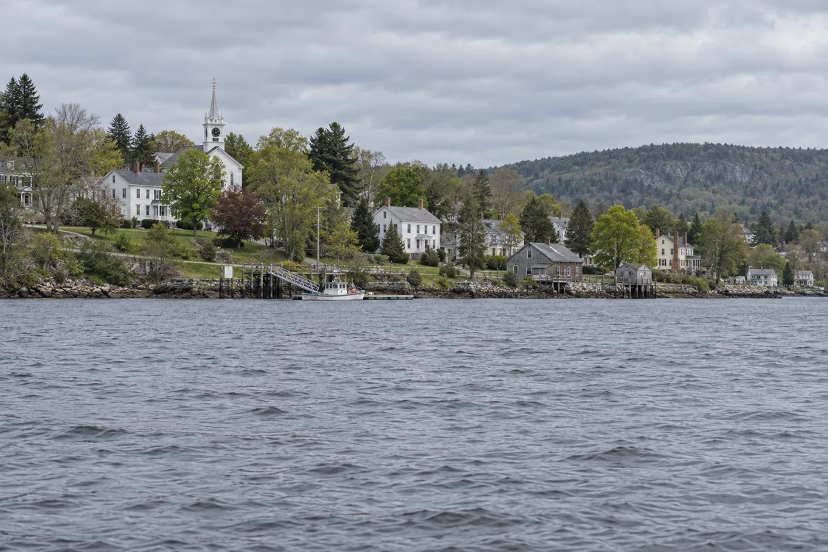 Winterport Maine village seen from the Penobscot River — white Greek Revival and Italianate captains' homes and a church steeple along Main Street rising from the west bank, the river wide and gray, the Bucksport hills across the water